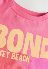 Tricou din bumbac roz, cu text mare galben care spune "SUNSET BEACH", având un model floral. Textură moale și design cu guler rotund.