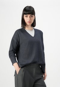 Maglione grigio scuro a scollo a V con maniche lunghe, vestibilità comoda e un trattamento a maglia leggermente testurizzato, indossato sopra una maglietta grigio chiaro. Pantaloni sartoriali a vita alta.