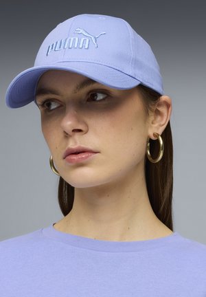 Mujer con una gorra Puma de color lila claro y un top a juego, lleva grandes pendientes de aro dorados, mirando ligeramente hacia su izquierda sobre un fondo gris.
