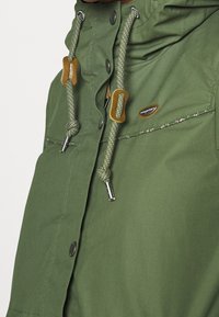 Grön vattentät jacka med dragsko i huvan, metalldragkedjor och tryckknappar. Tyget är slätt med en subtil glans.