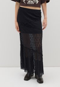 Schwarzer Spitzen-Maxikleid mit geschichteter Gestaltung, halbtransparenter Textur und elastischem Bund. Kombiniert mit dunklen Knöchelstiefeln.