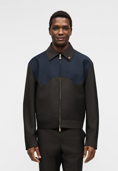 Άνδρας που φοράει μαύρο και μπλε ναυτικό μπουφάν με φερμουάρ και μακριά μανίκια, στέκεται μπροστά σε απλό ανοιχτό γκρι φόντο.