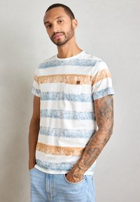 Mann med tatoveringer på høyre arm iført en hvit t-skjorte med bl�å og oransje striper og lyseblå jeans, poserer med hendene i lommene.