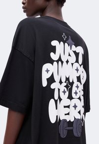 Άτομο που φοράει μαύρο t-shirt με μεγάλο λευκό κείμενο «Just pumped to be here» και γραφικό από αλτήρες στην πλάτη.