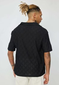 Chemise noire à manches courtes avec un motif de losanges texturé, dotée d'un col rabattable et d'une coupe décontractée, idéale pour une tenue décontractée.