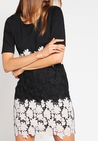 Robe noire à manches courtes en tissu lisse, avec un superposé en dentelle crème aux motifs floraux sur l'ourlet.