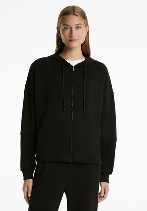 Eine Frau trägt einen schwarzen Reißverschluss-Hoodie mit Kordelzügen über einem weißen Oberteil, kombiniert mit schwarzen Hosen, und steht vor einem schlichten weißen Hintergrund.