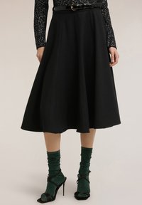 Sort midi-nederdel med udvidet design, struktureret tekstur og en linning med en rund bæltespænde. Stylet med sorte hæle med stropper og grønne sokker.