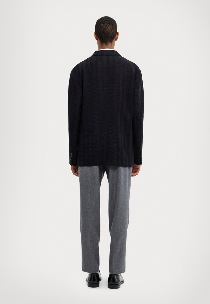 Blazer en chevron sombre avec un col cranté, associé à un pantalon gris clair. La tenue présente un design épuré et une coupe structurée.