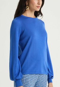 Pull-over tricoté bleu avec manches longues ballon et encolure ronde. Texture douce, poignets côtelés et coupe décontractée. Pantalon à motifs présenté en dessous.