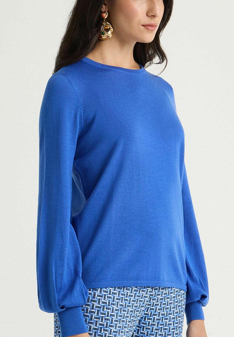 Pull-over tricoté bleu avec manches longues ballon et encolure ronde. Texture douce, poignets côtelés et coupe décontractée. Pantalon à motifs présenté en dessous.