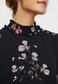 Blusa nera con stampa floreale in beige e rosa. Presenta un colletto arricciato ed elasticizzato. Tessuto leggero con una consistenza liscia.