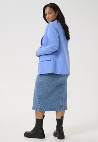 Blazer azul claro con un diseño ajustado, combinado con una falda de mezclilla hasta la rodilla y botas negras gruesas de tobillo, contra un fondo liso.