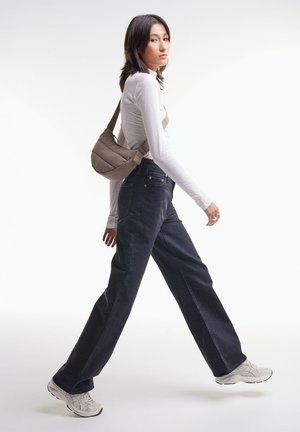 High-waisted dark denim jeans, white long-sleeve top, and light grey sneakers. The model carries a puffy beige shoulder bag with a curved design.