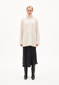 Gebreide trui in lichtbeige met een hoge geribbelde kraag, gecombineerd met een donkere gestreepte rok en kniehoge zwarte leren laarzen.