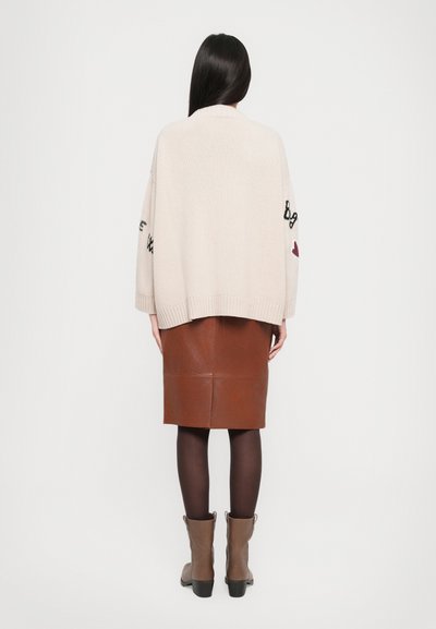 Cardigan in maglia crema con ricami neri e bordeaux, abbinato a una gonna in pelle marrone. Tessuto textured e design aderente, visibili stivali alla caviglia.