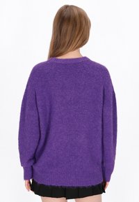 Pulover oversized, de culoare purpurie, cu o textură moale și pufoasă și manșete cu nervuri. Are un gât rotund și un tiv din spate ușor alungit.