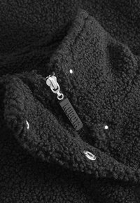 Vêtement noir en polaire avec un tissu texturé, fermeture éclair et étiquette de marque. Il présente des boutons-pression et des œillets métalliques pour un détail supplémentaire.
