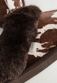 Braune Hausschuhe im Kuhflecken-Design mit weicher, flauschiger dunkelbrauner Innenseite und brauner Sohle. Mit einem Etikett mit der Aufschrift "NEXT". Genoppte Stoffoberfläche.