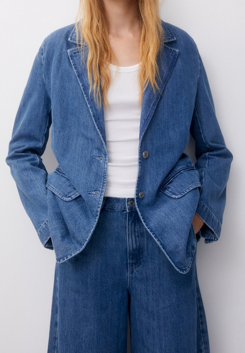 Femme portant un blazer en jean bleu et un jean à jambes larges assorties sur un débardeur blanc, les mains dans les poches du blazer.