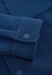 Prim-plan al unei mâneci din țesătură albastru marin, cu dungi striate și un nasture cu capse albastru asortat pe manșetă, evidențiind linii diagonale texturate.