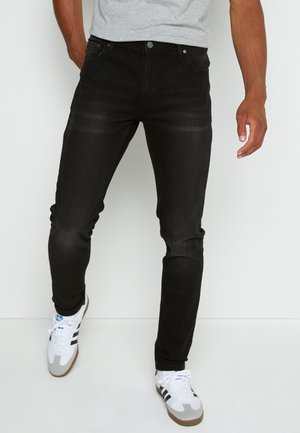 Mann in schmalen schwarzen Jeans und weißen Adidas-Sneakern, stehend vor einem weißen Hintergrund.