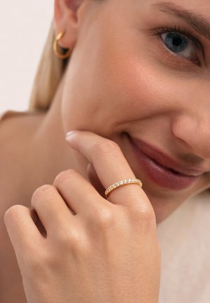 Anillo de oro con varias pequeñas piedras claras colocadas en fila; usado en una mano de piel clara, cerca de un rostro sonriente con maquillaje natural.
