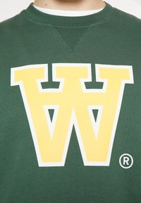 Zelená mikina s velkým žlutým logem "W" orámovaným bílou barvou, s kulatým detailem loga a žebrovým výstřihem.