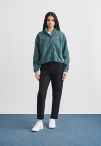 Naine seisab sinisel vaibal, kandes türkiissinist fliisjope, musti pükse ja valgeid tossusid, taustaks hall ühevärviline sein.