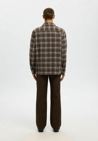 Vīrietis ar brūnu šļūtenes raksta garām piedurknēm kreklu un tumši brūnām biksēm, stāv ar muguru pret vienkāršu baltu fonu.