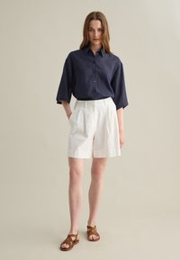 Navy linen button-up with short sleeves, paired with white pleated shorts and brown strappy sandals. Neutral background.