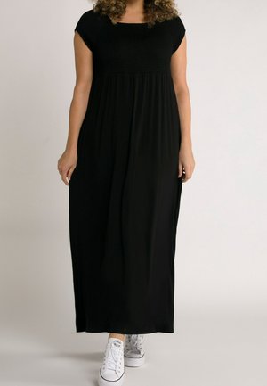 Vrouw draagt een lange zwarte jurk met korte mouwen, een gerimpeld lijfje en witte sneakers, staand tegen een effen lichte achtergrond.