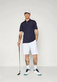 Ναυτικό πόλο μπλούζα, λευκά σορτς και λευκά αθλητικά παπούτσια με μπλε λεπτομέρειες. Κρατώντας ένα γκολφ κλαμπ, εμφανείς τατουάζ στα χέρια.