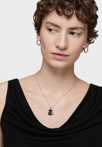 Giovane con capelli corti e ricci, che indossa orecchini a cerchio argentati e una maglietta nera, mentre guarda di lato, con una collana argentata che ha un piccolo ciondolo a forma di orsetto.