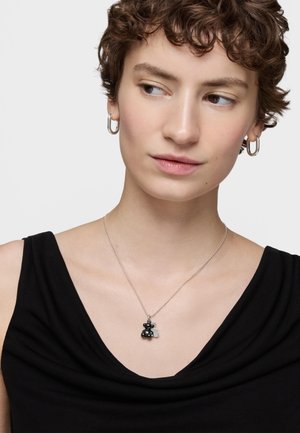 Joven con cabello corto y rizado, llevando aretes de aro plateados y una camiseta negra, mirando hacia un lado, con un collar plateado que tiene un pequeño colgante de oso.