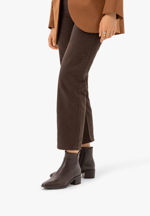 Personne portant des bottines marron, un pantalon marron foncé et une veste marron clair, debout avec une jambe légèrement avancée.