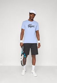 T-shirt azul claro de mangas curtas com uma estampa de um crocodilo preto, combinado com calções pretos e ténis brancos. A segurar uma raquete de ténis.