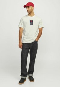Light gray short-sleeve t-shirt with a rectangular graphic print featuring a sun and wave pattern, paired with dark denim jeans and black shoes.