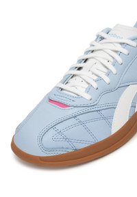 Šviesiai mėlyna odinė sportinė avalynė su baltais akcentais, turinti tekstūruotą paviršių, baltus raištelius ir rožinį akcentą netoli liežuvėlio. Ruda guma padas.