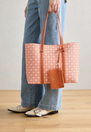Personne tenant un sac fourre-tout rose à motif avec des anses marron et un porte-cartes attaché, portant un jean bleu et des chaussures crème pointues avec des boucles.