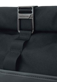 Svart ryggsäck med en texturerad tygdesign, som har ett metallspänne och en säkrad webband, vilket erbjuder en enkel och funktionell stängningsmekanism.