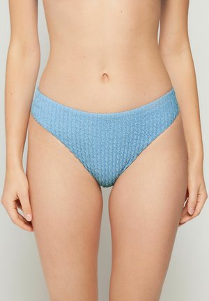 Frau trägt texturierte, hellblaue Bikinihose, steht vor einem einfarbigen hellen Hintergrund, die Hände locker an den Seiten.