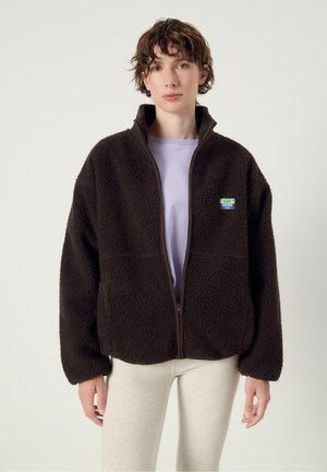 Junge Person mit kurzen braunen Haaren, die eine dunkelbraune Zip-Fleecejacke über einem lila Hemd und hellen Hosen vor einem einfarbigen Hintergrund trägt.