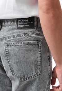 Jeans in denim grigio con finitura testurizzata, dotati di una tasca posteriore e di un'etichetta nera con testo bianco posizionata sulla cintura.