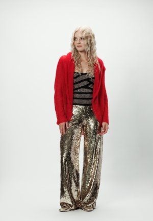 Kobieta o długich, kręconych blond włosach, ubrana w czerwony kardigan, czarny wzorzysty top oraz błyszczące złote spodnie o szerokich nogawkach, stoi na białym tle.