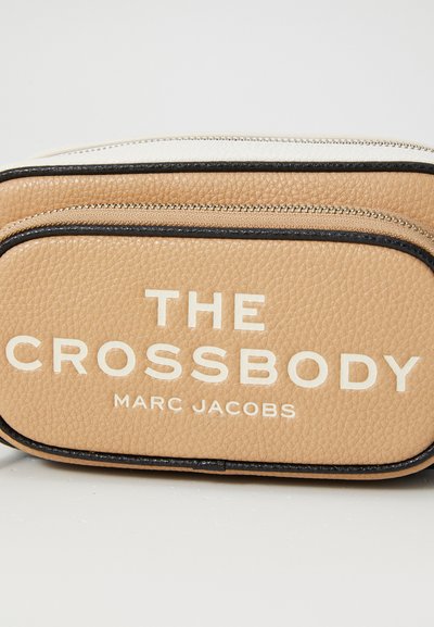 Tan leather crossbody bag with black accents, featuring a smooth texture, dual zip closures, and bold printed text: "THE CROSSBODY MARC JACOBS."