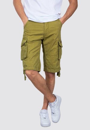 Mężczyzna w oliwkowo-zielonych szortach typu cargo i białych sneakersach, z rękami w kieszeniach, stoi ze skrzyżowanymi nogami na jednolitym tle.