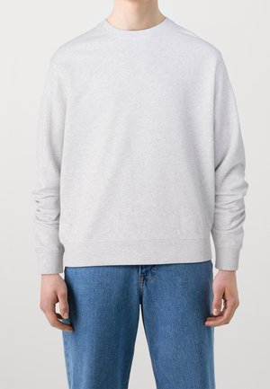 Persoon die een effen lichtgrijze sweater met lange mouwen en een blauwe spijkerbroek draagt, staand tegen een effen achtergrond.