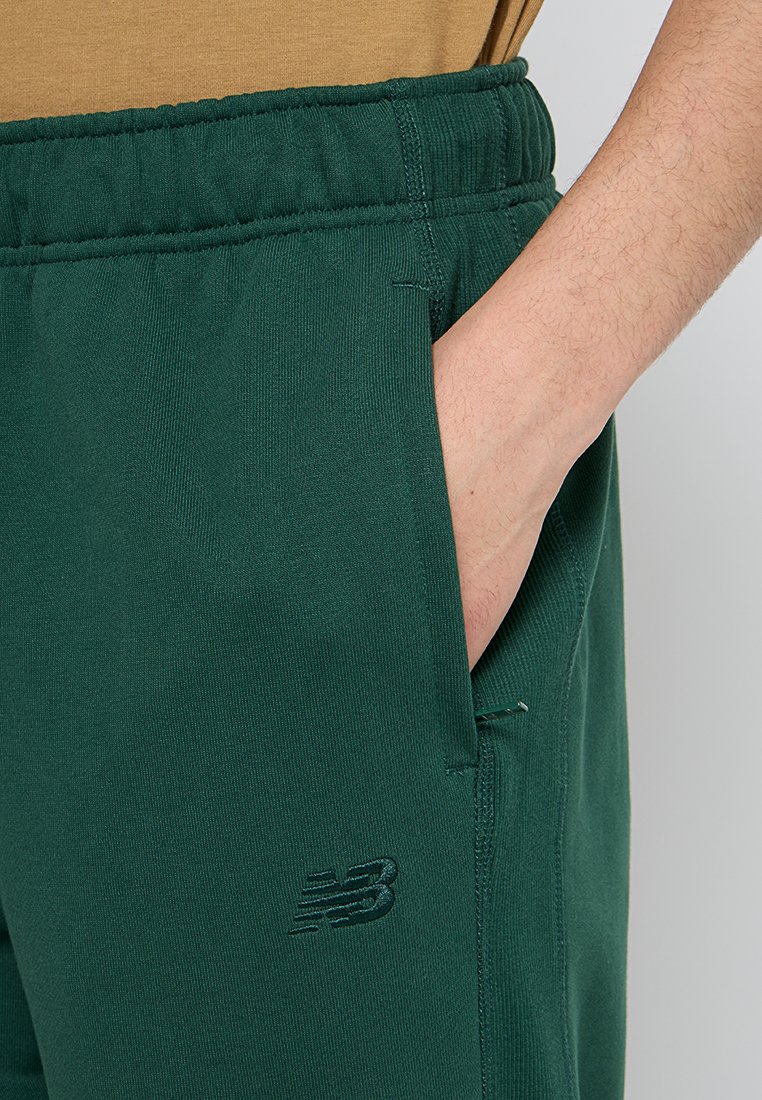 Pantaloni della tuta verdi con vita elasticizzata, tasche laterali e un sottile logo New Balance. Il materiale sembra morbido e ha una texture liscia.
