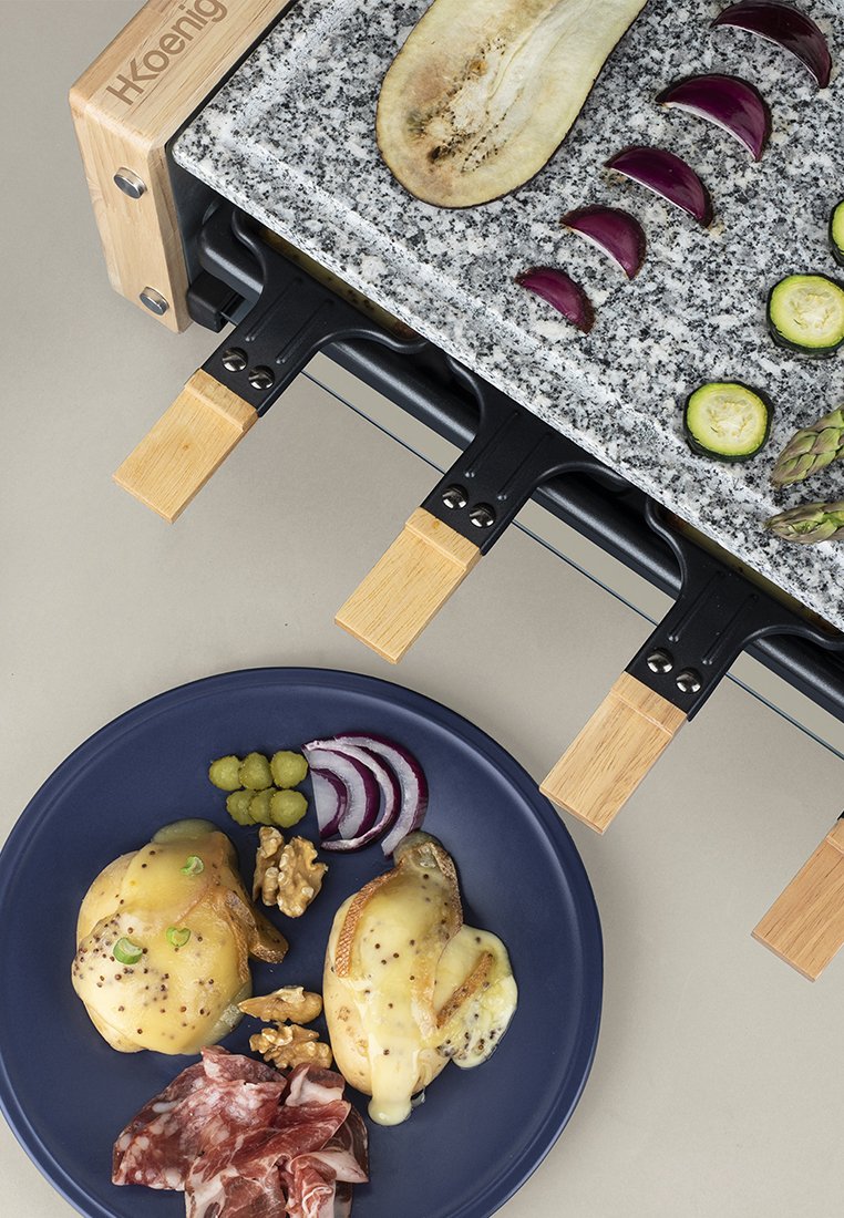 Gril à raclette avec une plaque en granit et des accents en bois. La plaque présente deux pommes de terre au fromage, des oignons rouges, des cornichons, des noix et de la viande séchée.